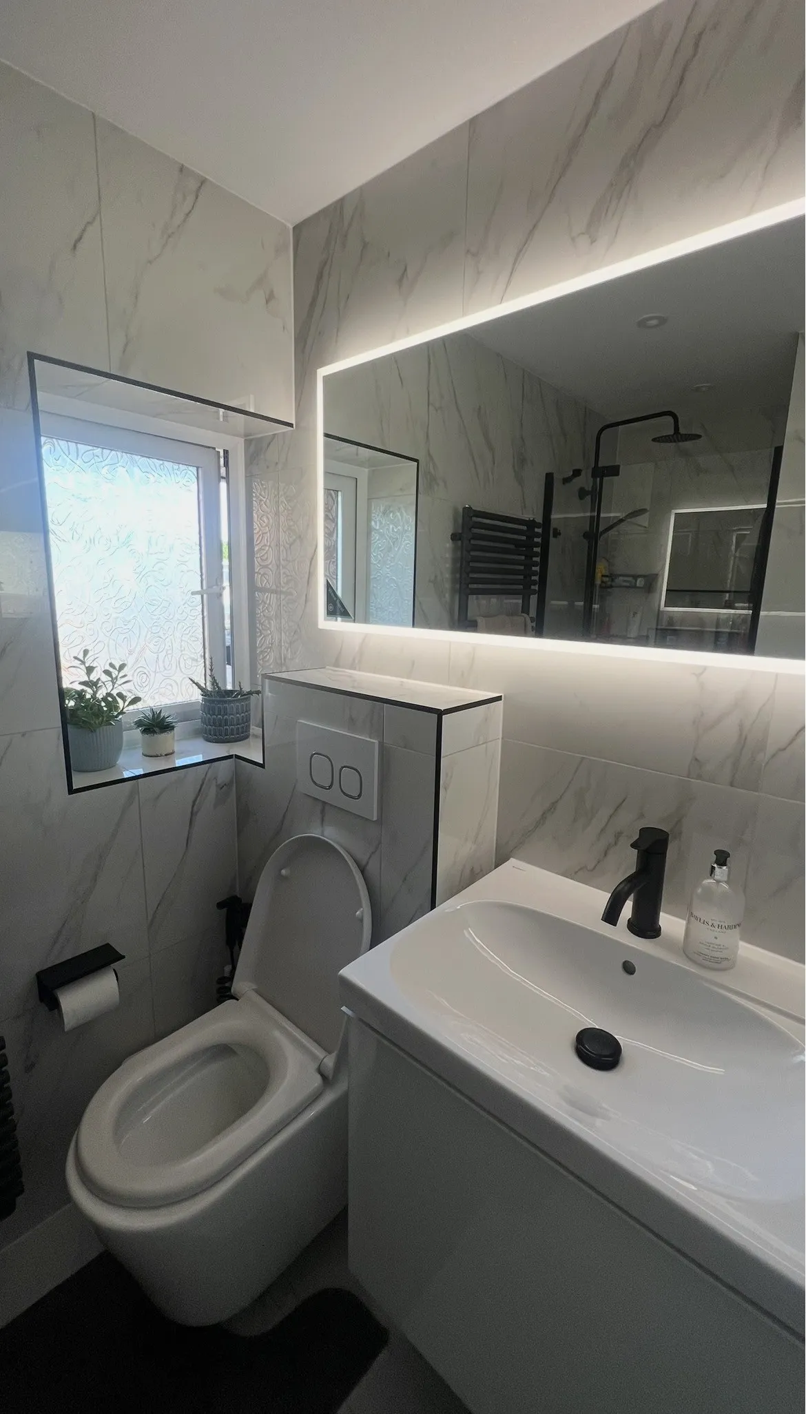 After: marble-effect porcelain walls, black-trimmed window reveal, wall-hung WC and backlit mirror